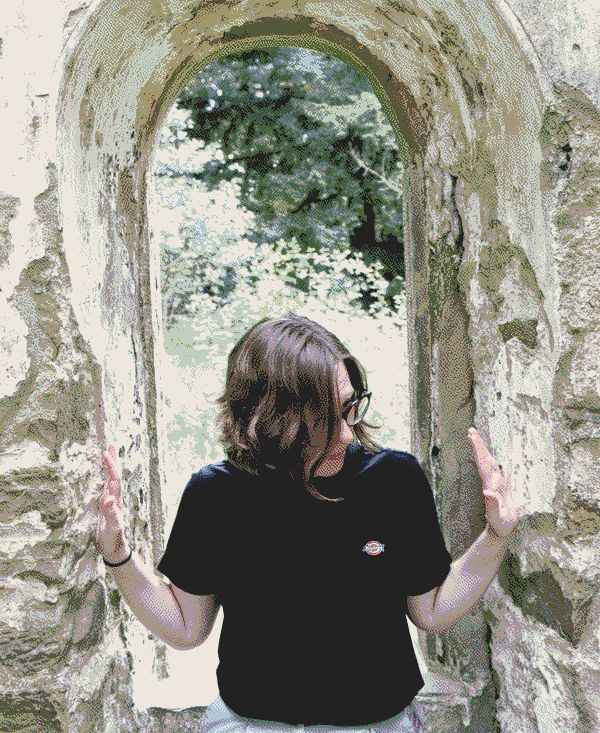 I am sitting in a stone arch window. I have my hands pressed gently on the stone on either side with my mid length hair covering my face. Through the window you can see lush green foliage.